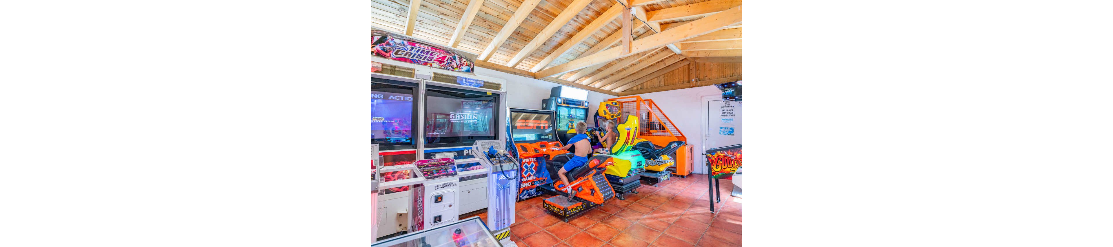 Two children sit on colorful motorcycle arcade machines in a bright indoor game room. Multiple large arcade cabinets and racing simulators line the tiled floor under a wooden ceiling. The space is filled with vivid screens, neon lights, and bold graphics. The scene captures a fun, energetic atmosphere of kids enjoying video games together.