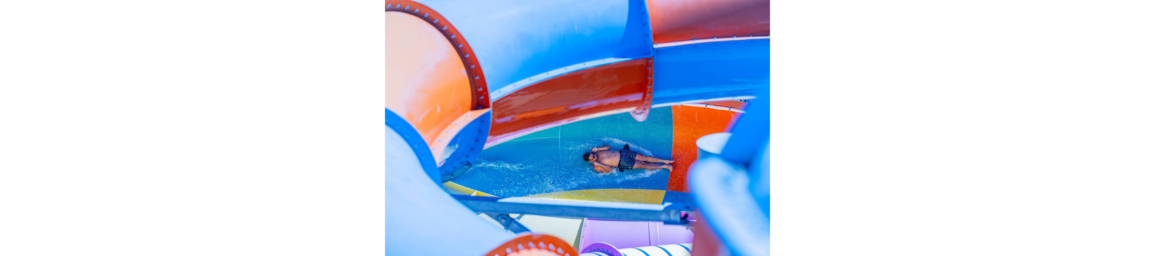 A man in swim trunks slides down a bright water slide at an outdoor water park. The scene is framed by large curved tubes in vivid blue, orange, and purple colors. Water splashes around him as he lies back and enjoys the ride. The overall image feels energetic, playful, and summery.