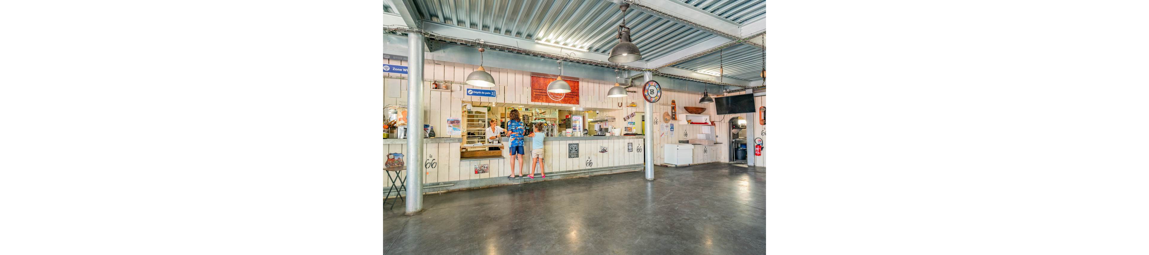 An indoor snack bar counter is set within a spacious industrial-style hall. Two children stand at the counter ordering food from a staff member inside the service window. The space features metal ceiling beams, hanging industrial lights, and polished concrete floors. White wooden wall panels are decorated with retro signs, Route 66 motifs, and a variety of small posters. A refrigerator, fire extinguisher, and other equipment line the back wall, adding to the casual, functional atmosphere.