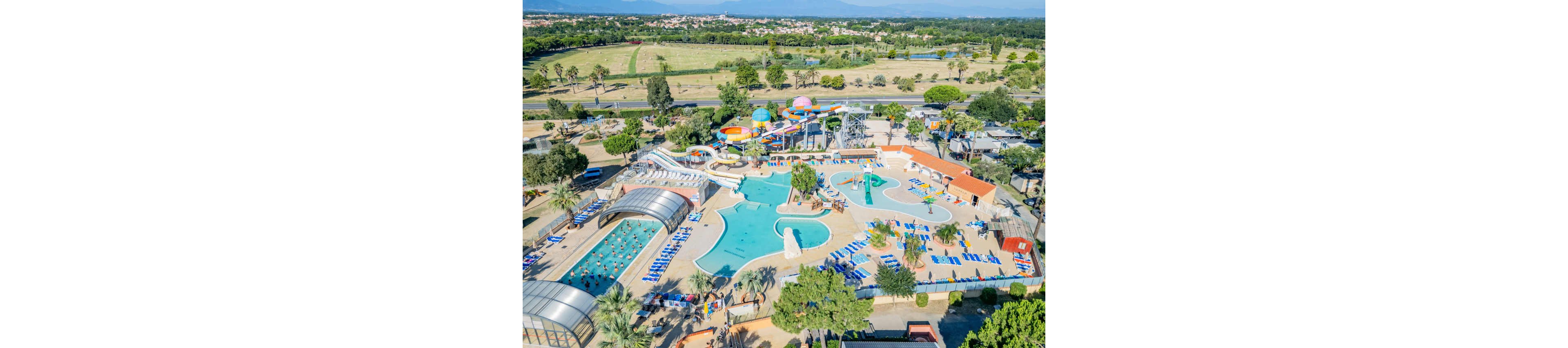 A large outdoor water park is captured from above, showing multiple pools, slides, and lounging areas. The complex includes a covered swimming pool, winding water slides, and a central splash zone with fountains. Numerous sun loungers are arranged around the pools, and colorful shade structures and play towers add visual interest. The park is surrounded by open fields, trees, and distant buildings, emphasizing its resort-like setting.