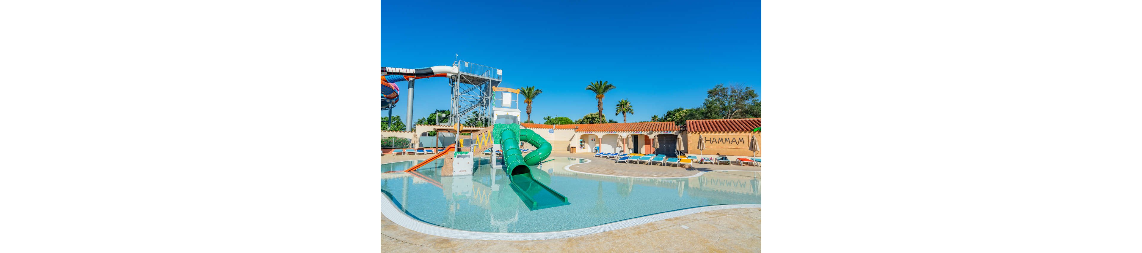 An outdoor resort pool features a bright green tube waterslide and a larger multicolored slide structure under a clear blue sky. The shallow pool curves around the slides, reflecting the surrounding buildings and palm trees. Sun loungers and umbrellas line the poolside, suggesting a relaxed vacation atmosphere. A building labeled "HAMMAM" is visible in the background, indicating spa facilities nearby.