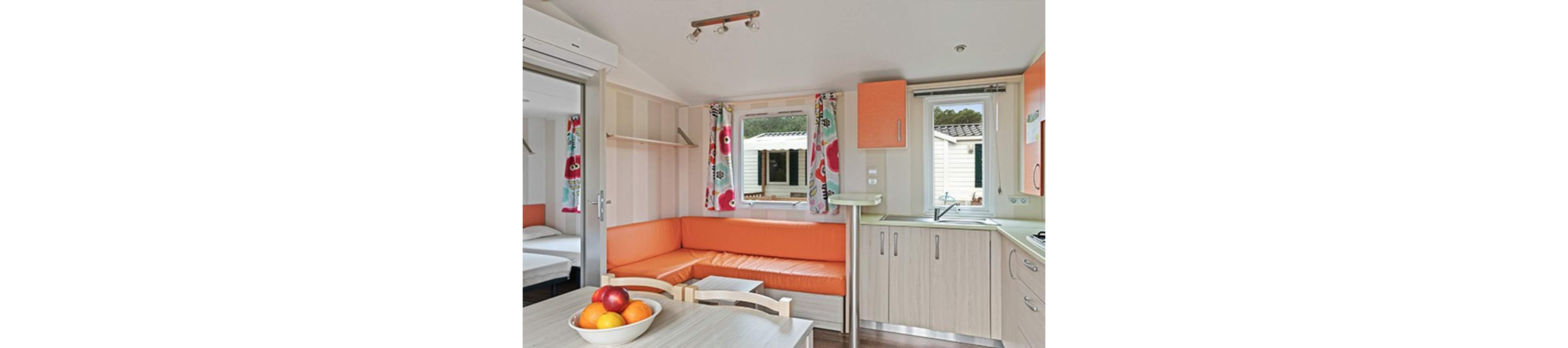 A compact holiday mobile home interior features a small kitchen and dining area with an orange corner sofa. Light wood cabinets and countertops frame a stainless steel sink and built-in cooktop. Colorful floral curtains and striped walls add a cheerful touch to the bright space. A bowl of fresh fruit sits on the dining table, and a bedroom with twin beds is visible through an open doorway.