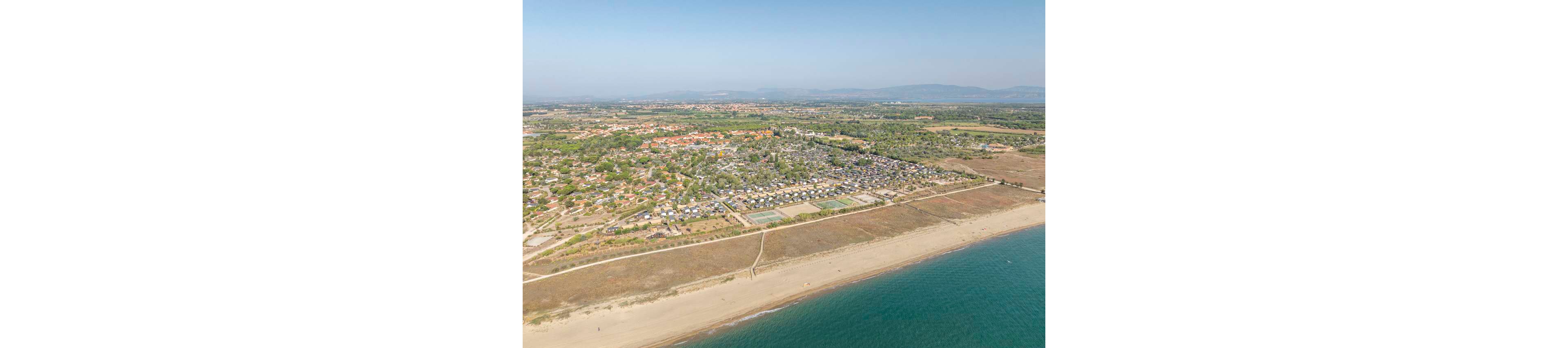 A wide aerial view shows a coastal holiday resort beside a long sandy beach. Rows of buildings, campsites, and recreational areas are visible behind the shoreline. Tennis courts and organized plots suggest a vacation village layout. The calm blue sea borders the beach, while green fields and distant hills stretch into the background under a clear sky.