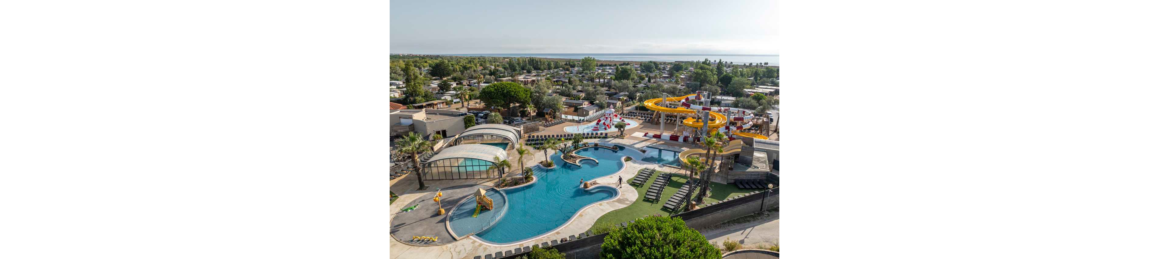 A large resort-style water park is captured from an elevated aerial perspective. Multiple outdoor swimming pools with curved shapes, slides, and splash structures are surrounded by sun loungers and palm trees. Bright yellow water slides and a covered pool area stand out as key visual elements. In the background, a campground or holiday park with numerous trees and buildings extends toward the distant coastline under a clear sky.