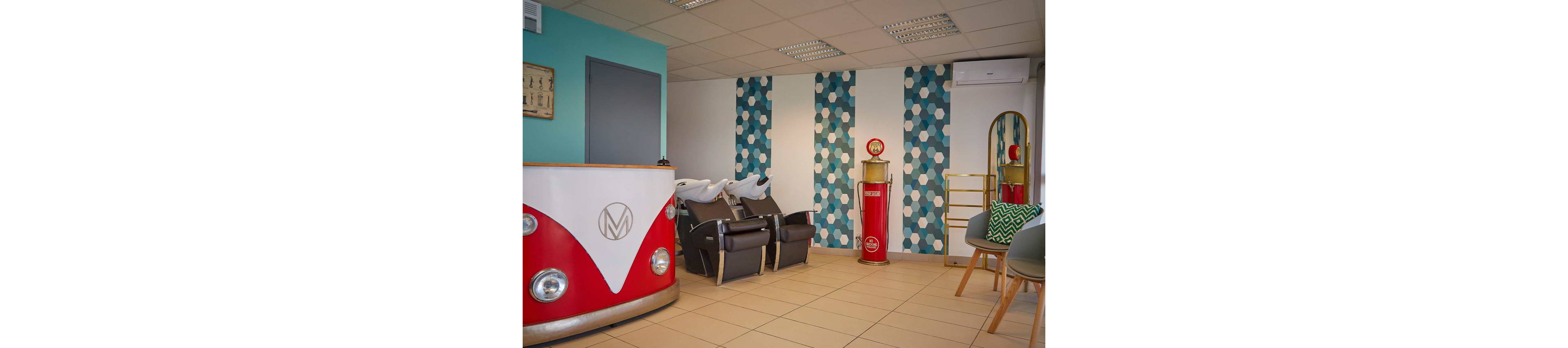 Colorful hair salon interior featuring a retro-style front desk shaped like a classic van. Two black shampoo chairs with white wash basins are positioned against a wall with blue and white hexagonal patterned panels. A red vintage-inspired column and decorative mirror stand add to the nostalgic theme. The room includes tiled flooring, modern ceiling lights, and a small seating area with green and patterned cushions.