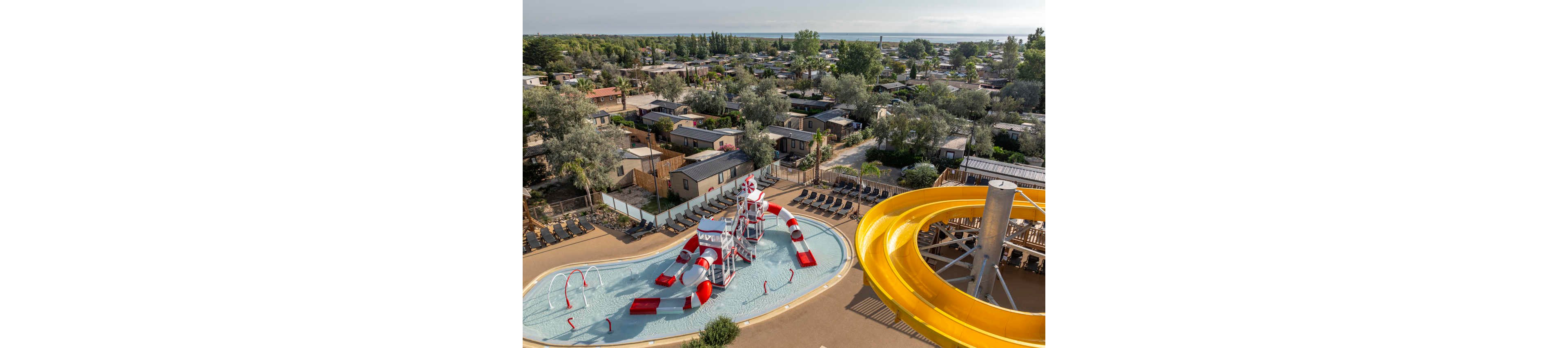 A colorful water play area with slides and climbing structures sits at the center of a holiday campsite. The scene includes a shallow pool with red and white play equipment and a large yellow water slide in the foreground. Rows of lounge chairs surround the pool, while mobile homes and cabins extend into the distance. Trees and greenery frame the resort, with a body of water visible on the horizon under a clear sky.