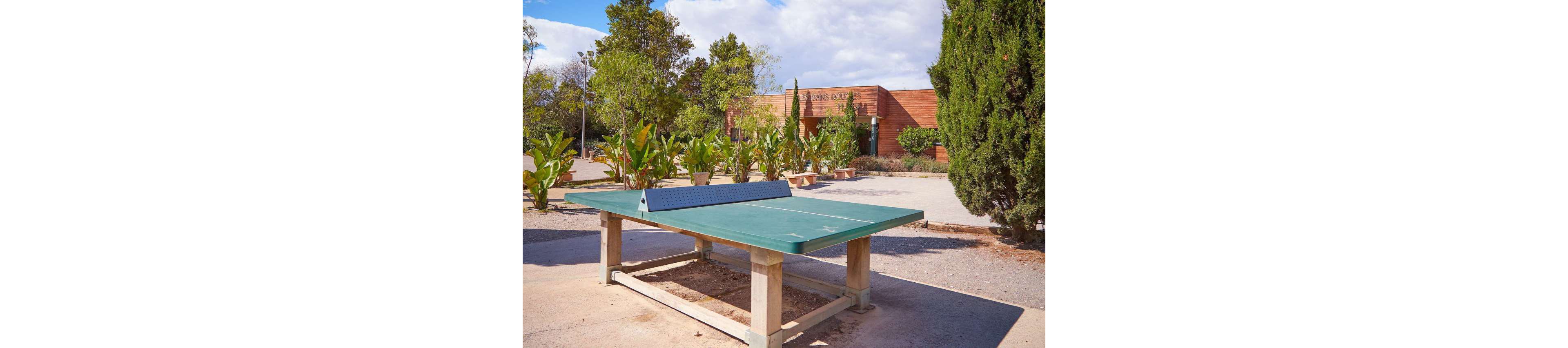 A green outdoor ping pong table stands in an open courtyard under a bright sky. The sturdy table is mounted on a concrete base with a metal net in the center. Surrounding the area are potted plants and trees, adding greenery to the scene. In the background, a low modern building with wooden cladding is partially visible.