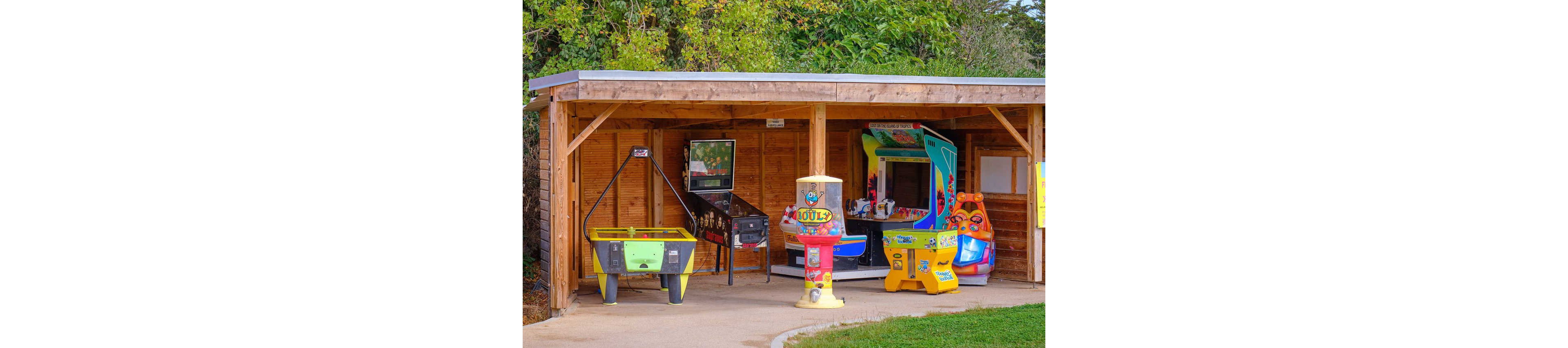 A small wooden shelter houses several colorful arcade and amusement machines. Visible games include an air hockey table, a pinball machine, a claw crane, and kiddie ride machines. The shelter is set in an outdoor park or recreation area with greenery in the background. Bright colors and playful graphics create a fun, family-friendly atmosphere.