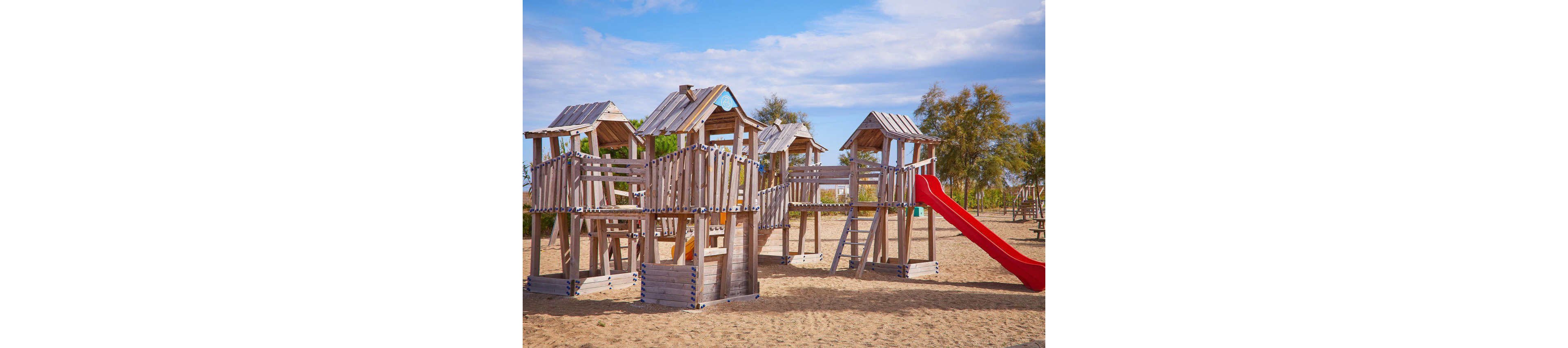 A large wooden playground structure stands on a sandy beach under a partly cloudy sky. Multiple towers, bridges, and ladders create an adventurous play area for children. A bright red slide adds a vivid accent to the otherwise natural wood tones. Trees and sparse vegetation appear in the background, suggesting a coastal recreational area.