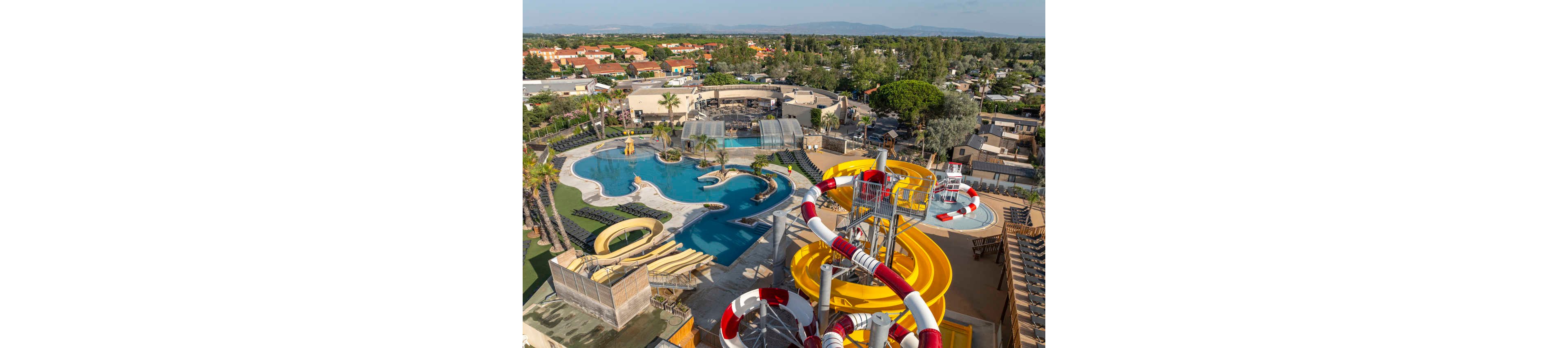 A large resort water park features multiple twisting water slides and a freeform swimming pool. The scene is captured from above, showing bright yellow and red slides spiraling toward splash pools. Sun loungers and palm trees surround the pool area, with covered seating and landscaped walkways visible. In the background, residential buildings and greenery stretch toward distant hills under a clear blue sky.