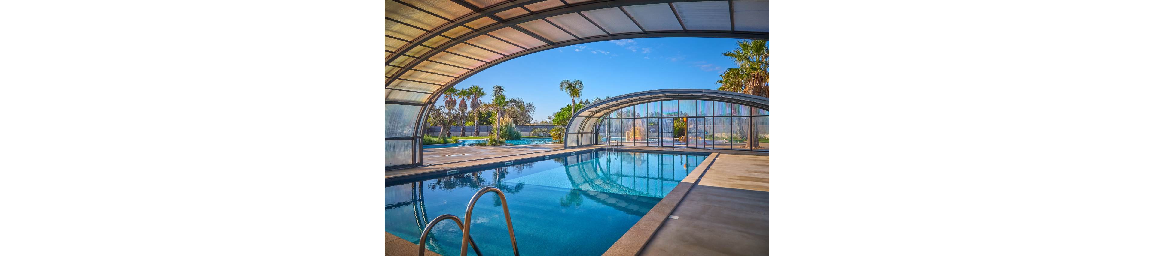 A clear blue swimming pool sits beneath a modern curved glass enclosure. The structure opens to an outdoor area with palm trees and a bright blue sky. Sunlight reflects off the calm water, creating a serene and inviting atmosphere. Metal pool ladders and wide concrete decking frame the pool, suggesting a resort or private leisure setting.