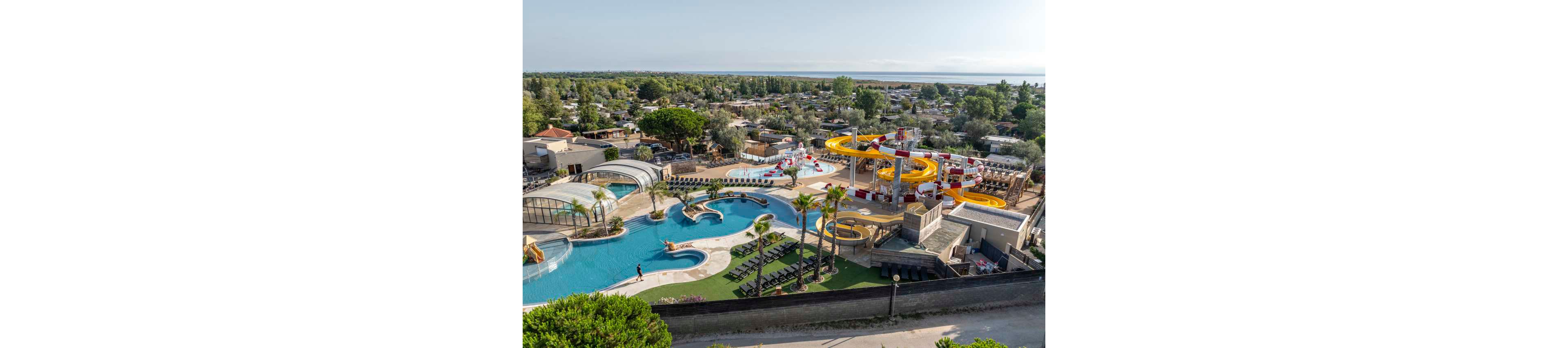 A large outdoor waterpark and swimming pool complex is seen from above within a holiday resort. Curving yellow waterslides, splash play structures, and multiple pools dominate the scene. Sun loungers and palm trees surround the water areas, creating a relaxing vacation atmosphere. In the background, numerous cabins and trees stretch toward the horizon under a clear sky.