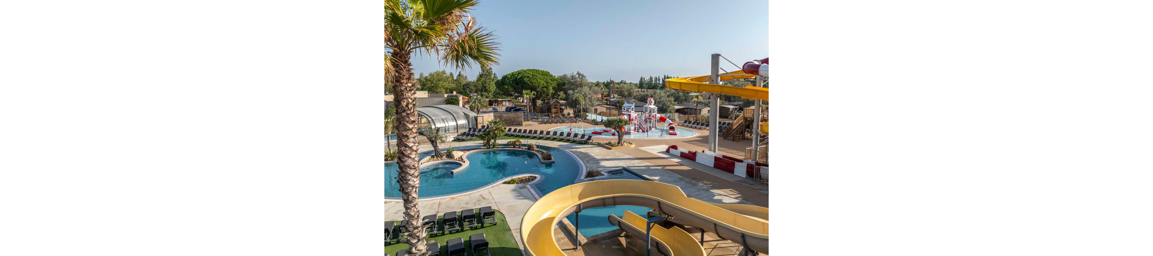 An expansive outdoor water park features winding water slides, curved swimming pools, and a dedicated children's splash area. Lounge chairs line the poolside terraces, offering plenty of space for sunbathing and relaxation. Palm trees and landscaped greenery add a tropical feel to the resort-like setting. Clear skies and bright sunlight create a warm, inviting atmosphere for families and vacationers.