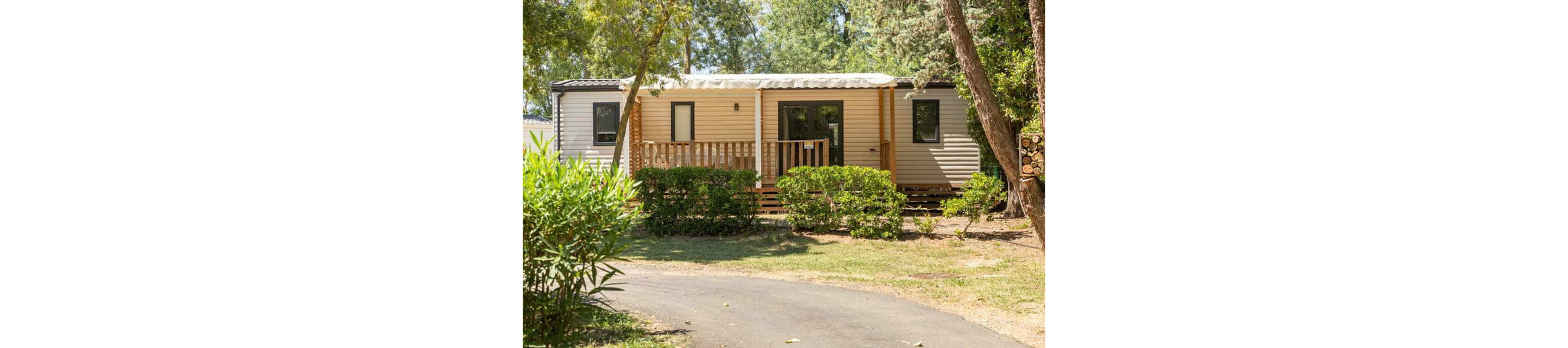A beige mobile home with a small wooden deck is set among lush green bushes and tall trees. The structure features several windows and a central glass door opening onto the porch. A paved path curves in front of the home, leading through the shaded campsite area. Bright daylight and abundant foliage create a peaceful, vacation-like atmosphere.