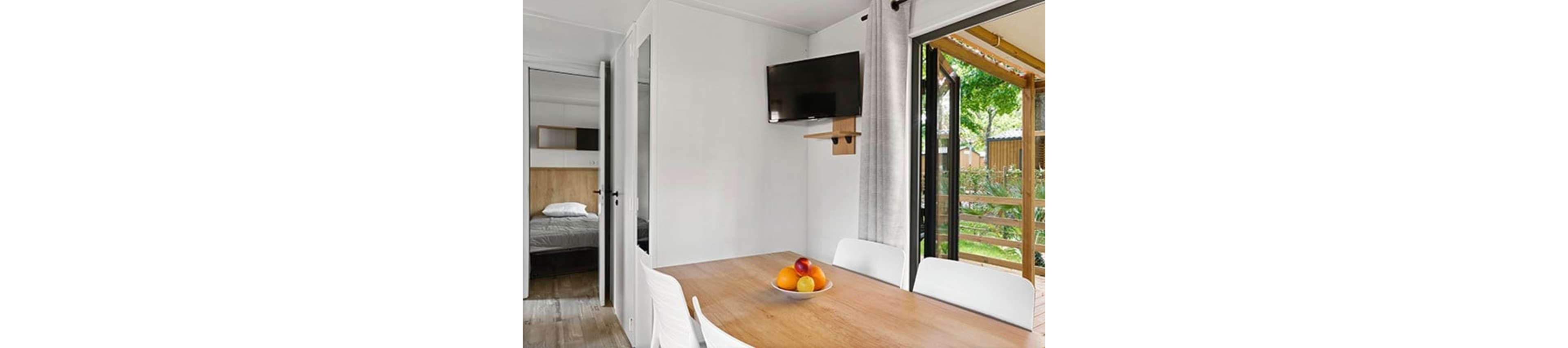 A compact dining area with a wooden table and white chairs is shown inside a modern mobile home. A bowl of colorful fruit sits on the table, adding a fresh accent to the minimalist decor. A flat-screen television is mounted on the wall above light gray curtains. An open sliding door reveals a small terrace with wooden railing and greenery outside, while a bedroom with a neatly made bed is visible through an interior doorway.