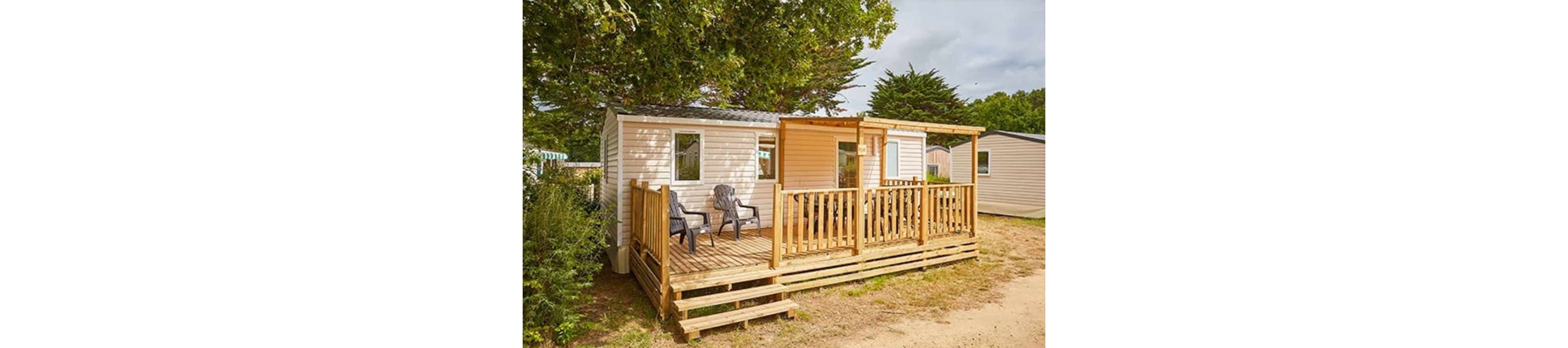 A compact mobile home features a spacious wooden deck with railings and steps leading down to a sandy ground. Two plastic chairs sit on the terrace, suggesting a relaxed outdoor seating area. The home is surrounded by trees and greenery, creating a peaceful, natural setting. Neighboring mobile homes are visible in the background, indicating a holiday park or campground environment.