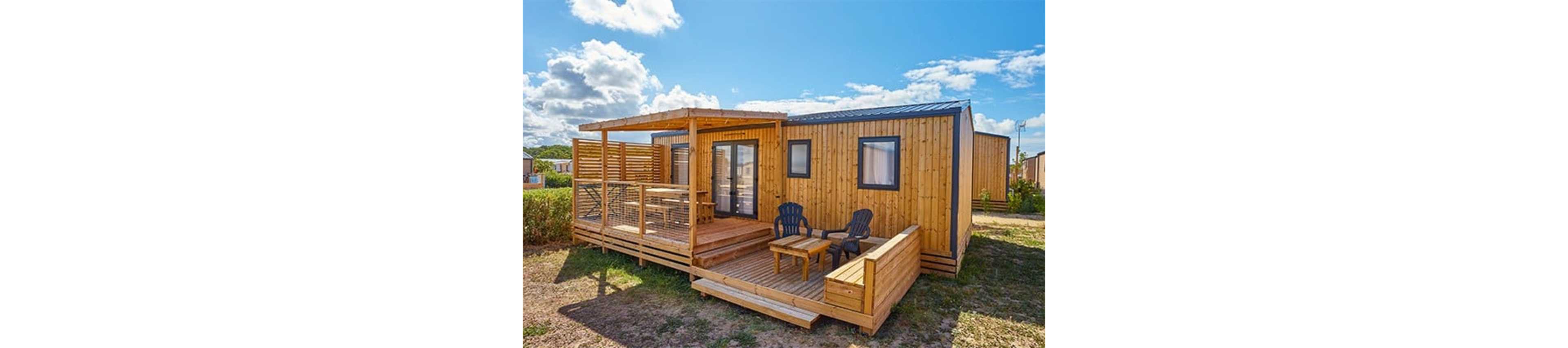 A compact wooden tiny house stands in an open outdoor setting under a bright blue sky with scattered clouds. The structure features a spacious front deck with a covered pergola area and railing. Two dark plastic chairs and a small table are arranged on the uncovered part of the deck, suggesting a relaxing seating area. The surrounding landscape appears dry and grassy, with hedges and other small structures visible in the background.