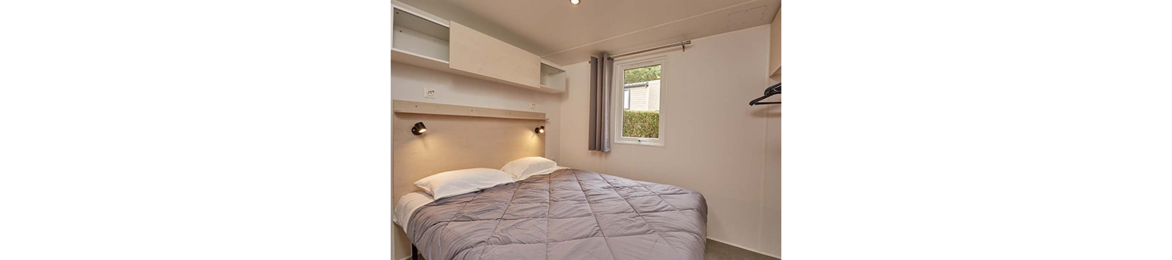 A compact bedroom features a neatly made double bed with a gray quilt and white pillows. Built-in shelves and a light wood headboard with two reading lamps create a minimalist, functional look. A small window with gray curtains lets in natural light and offers a view of greenery outside. The neutral color palette and simple furnishings give the room a calm, modern atmosphere.
