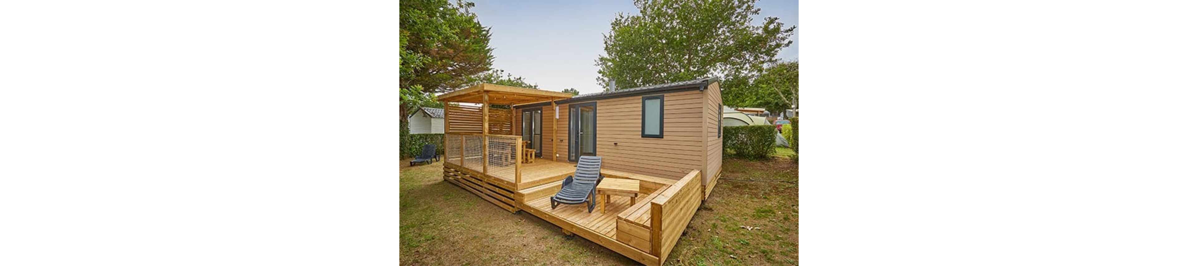 A contemporary wooden mobile home features a spacious raised terrace in an outdoor setting. The deck includes a pergola-covered dining area with chairs and an open section with a lounge chair and small table. Large glass doors connect the interior to the terrace, enhancing indoor-outdoor living. The home is surrounded by grass, hedges, and mature trees, suggesting a quiet campsite or holiday park environment.
