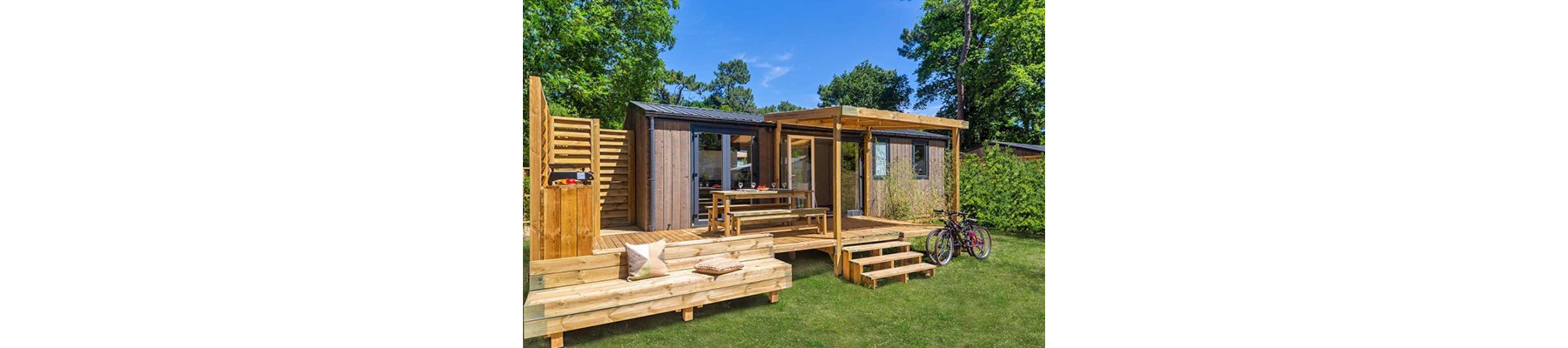 A contemporary wooden tiny house sits in a lush green yard surrounded by trees. The structure features large windows, a covered terrace with a dining table, and an upper deck accessed by exterior stairs. Wooden steps and benches extend into the lawn, creating an inviting outdoor living area. Two bicycles are parked near the porch, suggesting leisure and outdoor activities.