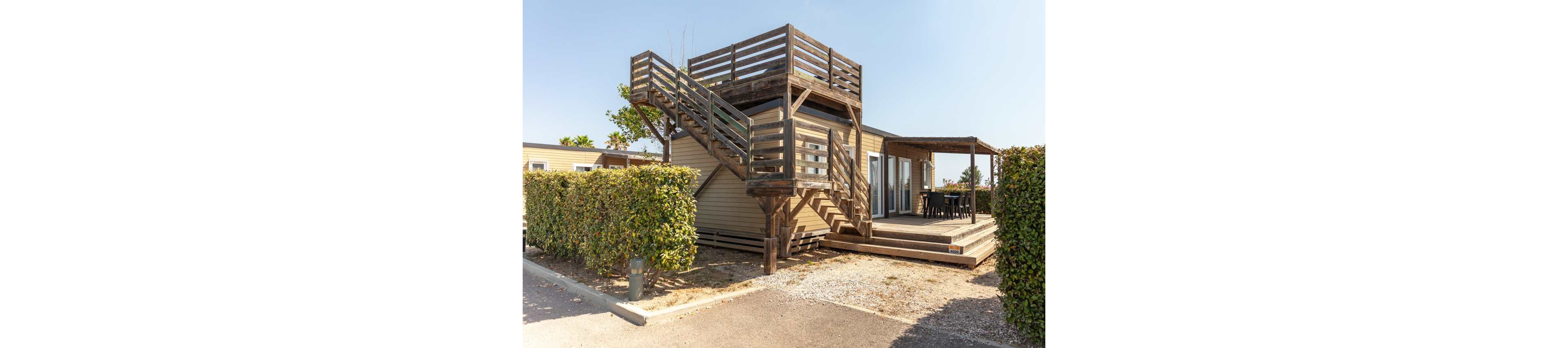 A contemporary wooden cabin features an elevated rooftop terrace accessed by an exterior staircase. The structure sits in a sunny outdoor setting with trimmed hedges and a gravel driveway. Large glass doors open onto a spacious ground-level deck with an outdoor dining table under a pergola. The scene suggests a relaxed vacation rental or holiday home in a warm climate.