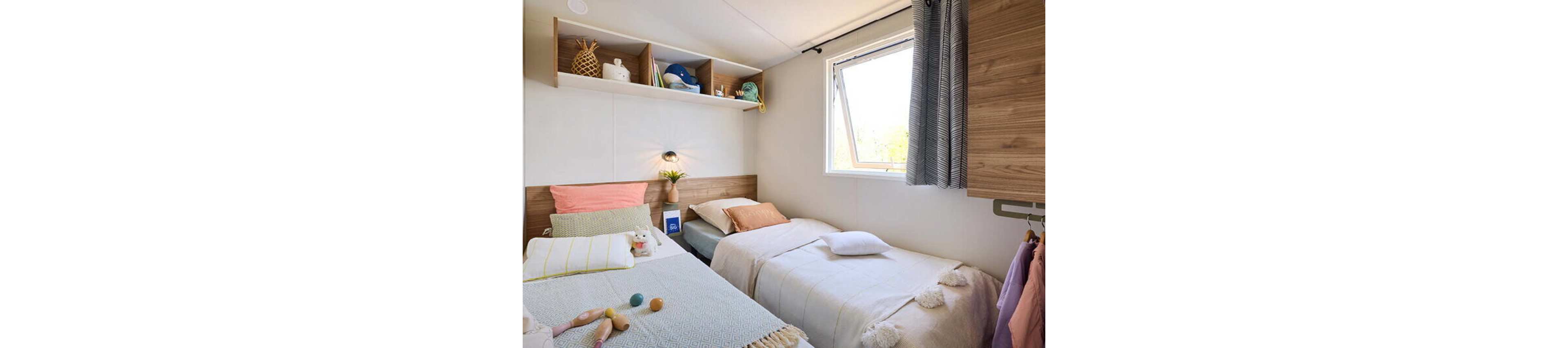 Small modern bedroom featuring two neatly made single beds with soft pastel bedding. The room includes wooden accents, a wall-mounted reading light, and open shelving displaying decor items and toys. A window lets in natural daylight, illuminating the neutral walls and light curtains. Clothes hang on a rail to the side, adding a lived-in yet tidy feel to the space.