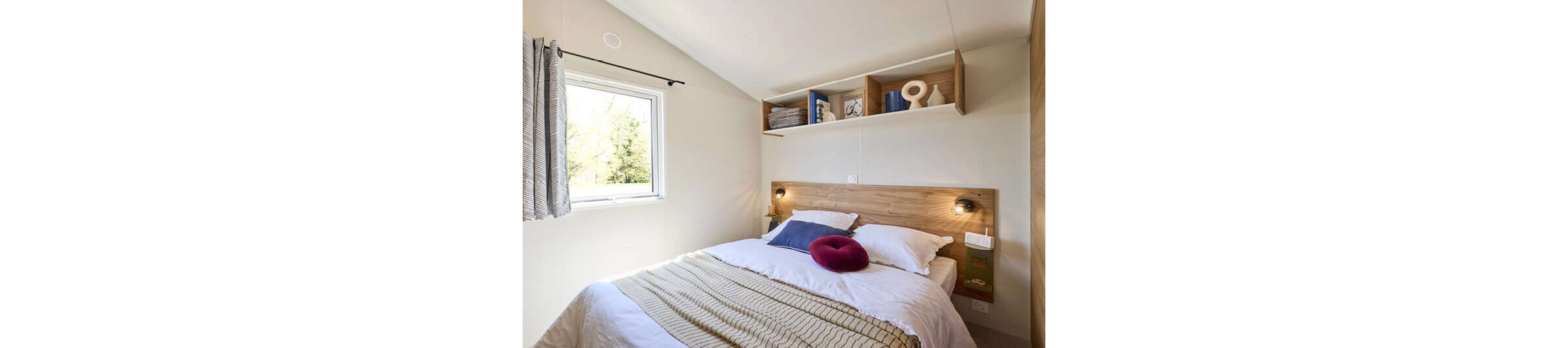 A compact bedroom features a neatly made double bed with white linens and a beige knitted throw. A round red cushion adds a pop of color against the neutral bedding. Above the bed, wooden shelves hold folded towels, books, and decorative objects. Natural light enters through a window with striped curtains, creating a bright and inviting atmosphere.