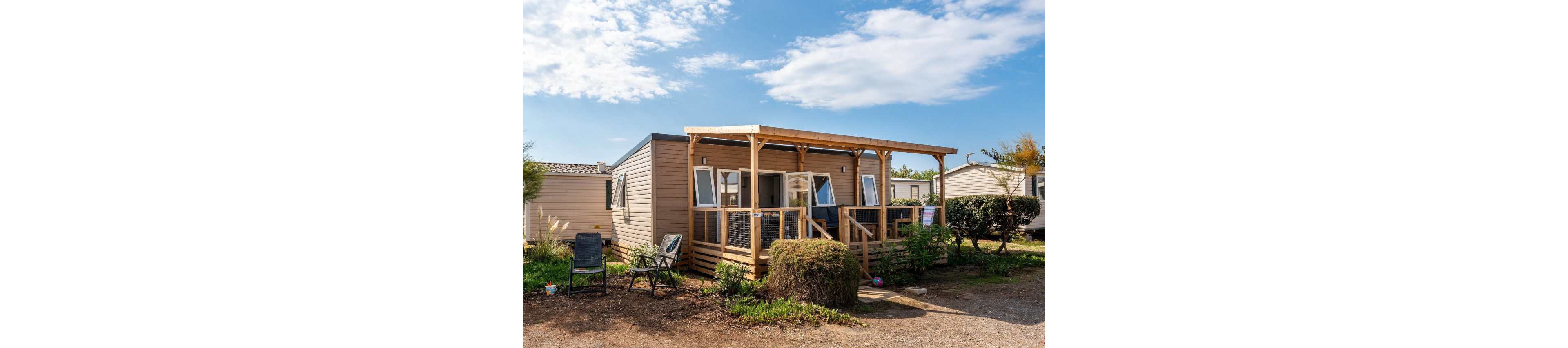 A modern mobile home with a covered wooden terrace stands in a holiday park under a bright blue sky. Outdoor chairs and a small table are arranged in front, suggesting a relaxing seating area. The structure features large windows and sliding doors that open onto the deck, surrounded by low shrubs and neighboring units. The overall scene conveys a peaceful vacation setting in a well-kept campground.
