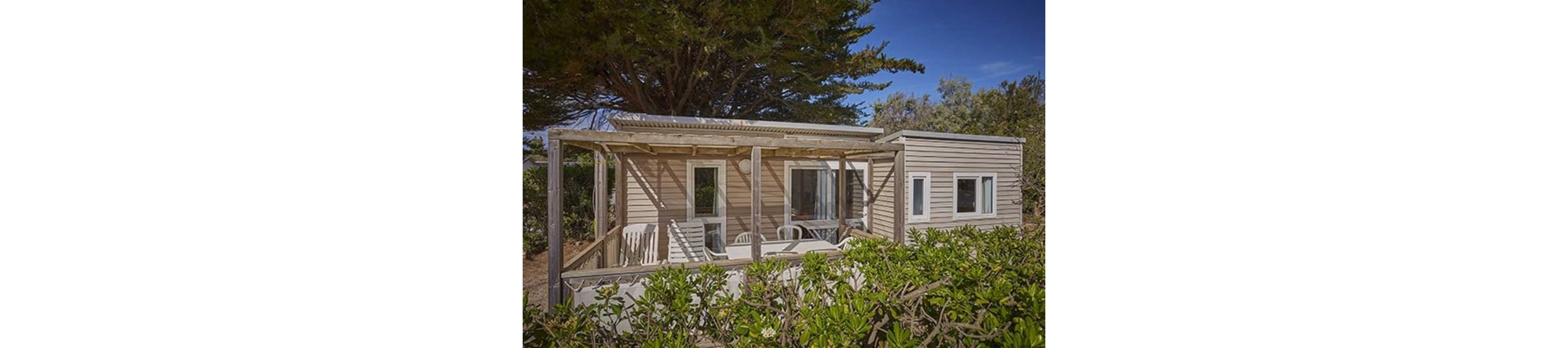 A small wooden cabin with a covered terrace is nestled among dense green shrubs and trees. The structure features large windows and a simple pergola-style roof over an outdoor seating area with white plastic chairs and a table. Bright daylight and a clear blue sky suggest a warm, pleasant climate. The scene conveys a quiet, relaxed vacation or countryside retreat atmosphere.