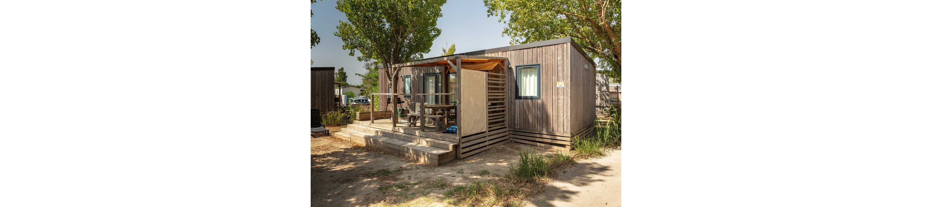 A compact wooden cabin stands in a sunny outdoor setting surrounded by leafy trees. The structure features a small raised deck with railings and a partially covered terrace area. Outdoor furniture is visible under the shade, suggesting a space for relaxing or dining. The ground around the cabin is dry and sandy, evoking a warm, summer holiday atmosphere in a campground or vacation park.