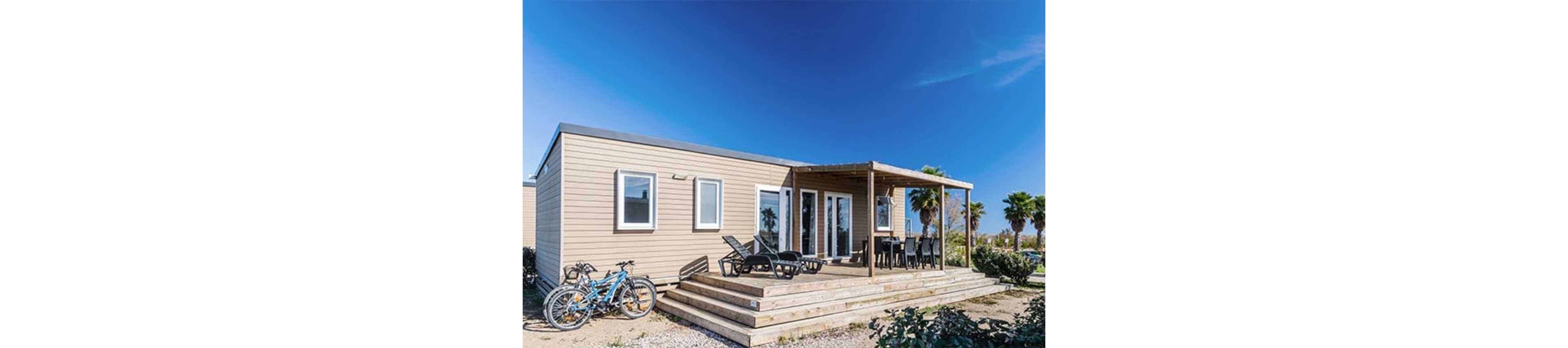 A contemporary mobile home with light wood siding stands under a clear blue sky. The front features a spacious wooden deck with lounge chairs and a shaded pergola dining area. Two bicycles are parked near the steps, suggesting outdoor recreation. Palm trees and low vegetation surround the home, giving the scene a relaxed vacation atmosphere.