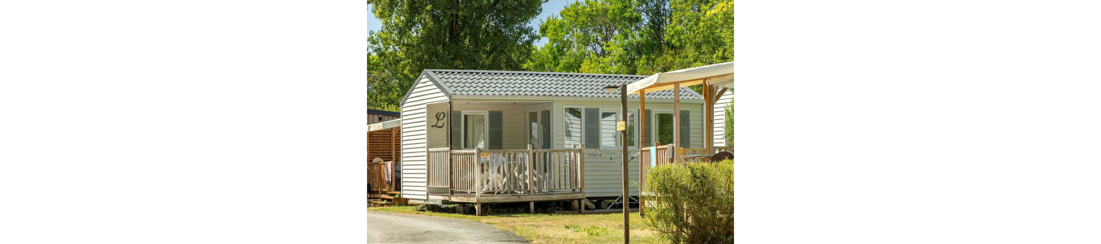 A white mobile home with a small wooden porch stands in a sunny campsite. Several white plastic chairs are arranged on the deck, suggesting a relaxed outdoor seating area. The cabin is surrounded by green trees and shrubs, with a gravel path running in front. Warm daylight and clear weather create a peaceful holiday atmosphere.