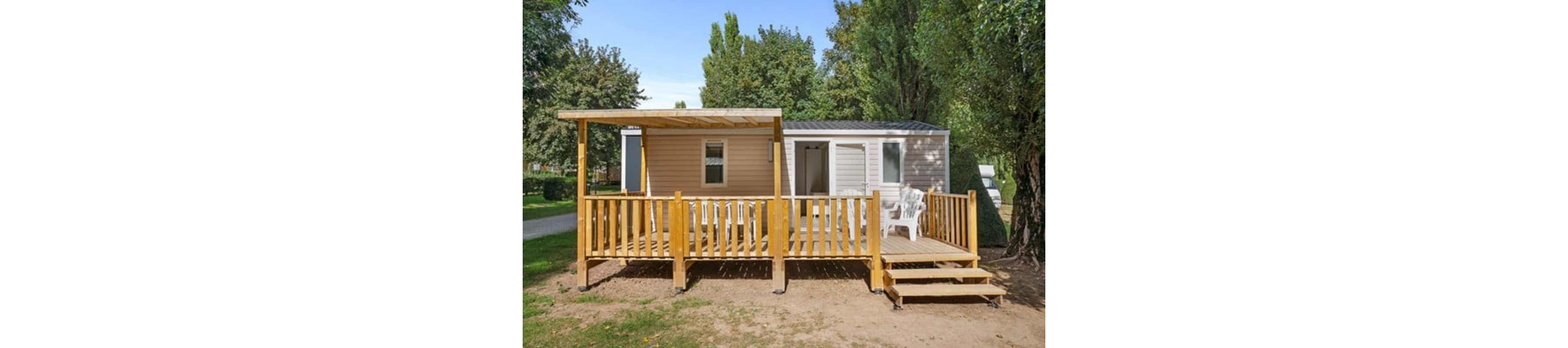 A compact mobile home features a spacious wooden terrace with railing and steps leading up from the ground. The structure is set in an outdoor campground or holiday park surrounded by tall green trees. White plastic chairs sit on the deck, suggesting a relaxed outdoor seating area. The scene is bright and clear, emphasizing leisure and vacation living in a natural setting.