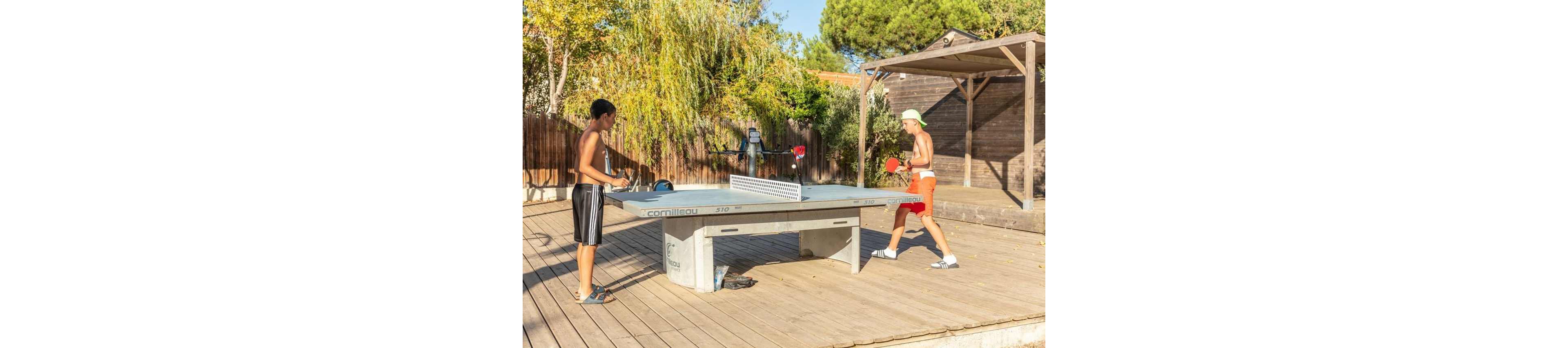 Two young men play an intense game of table tennis on a wooden deck. The match takes place outdoors in a sunny setting with trees, a wooden fence, and a pergola in the background. Both players wear casual summer clothing, including shorts and sandals, and hold red paddles. The bright daylight and warm tones create a relaxed, vacation-like atmosphere.