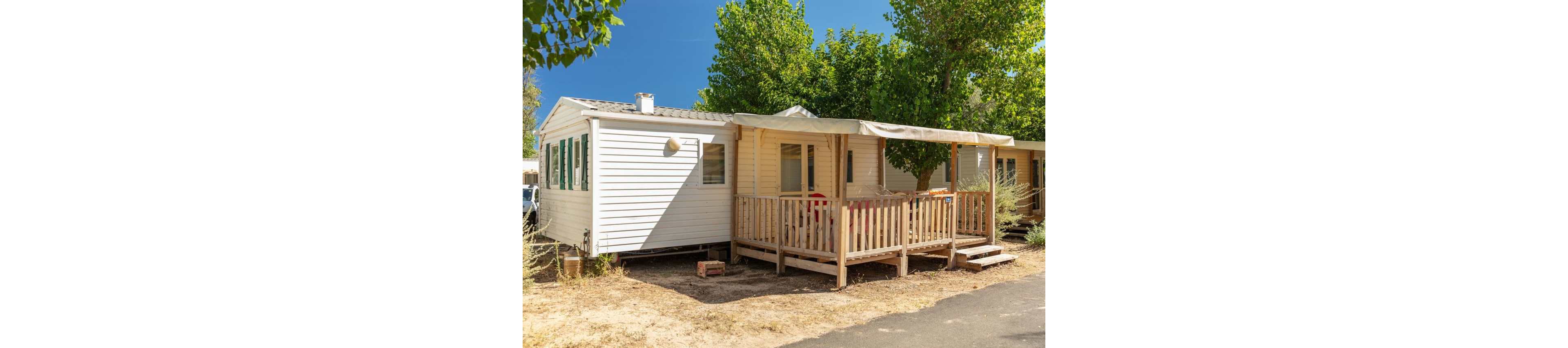 A white mobile home with a small wooden terrace stands in a sunny outdoor setting. The terrace is covered with a light canopy, providing shade over the entrance area. Surrounding trees offer additional greenery and contrast with the dry ground and paved path in front. The scene suggests a relaxed vacation or campsite environment in warm weather.