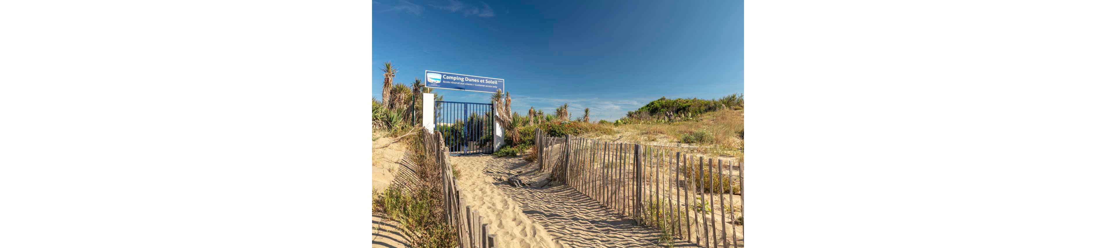 A sandy path between wooden fences leads to a closed metal gate at the entrance of a campsite. The sign above the gate reads "Camping Dunes et Soleil" against a clear blue sky. Low vegetation and dune grasses line the pathway, suggesting a coastal environment. Strong sunlight creates long shadows and emphasizes the warm tones of the sand.