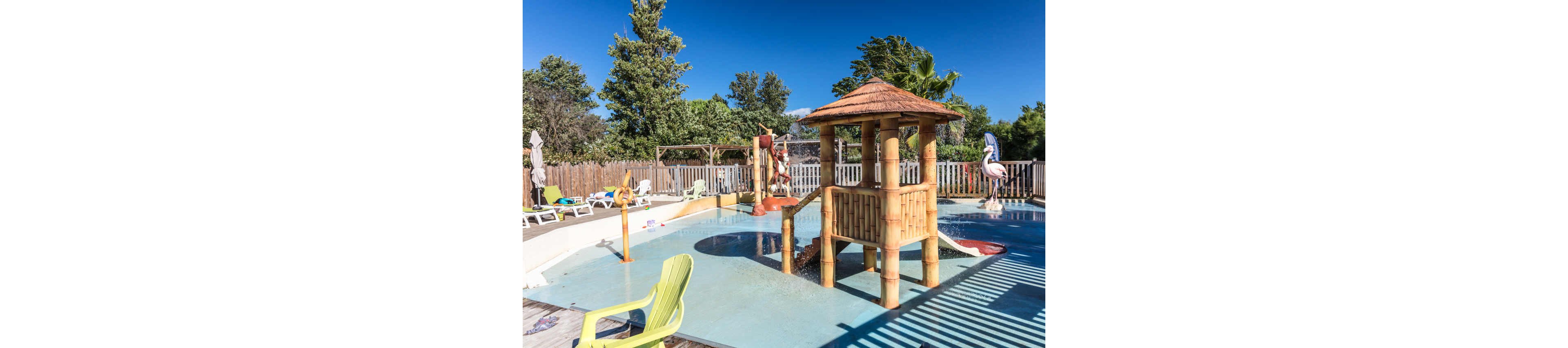 An outdoor shallow splash pool features a wooden play structure with a small slide and water features. The pool is surrounded by lounge chairs and a fence, set under a clear blue sky. Tropical-style elements, such as bamboo posts and a thatched roof, create a relaxed vacation atmosphere. Trees and greenery in the background add to the resort-like setting.