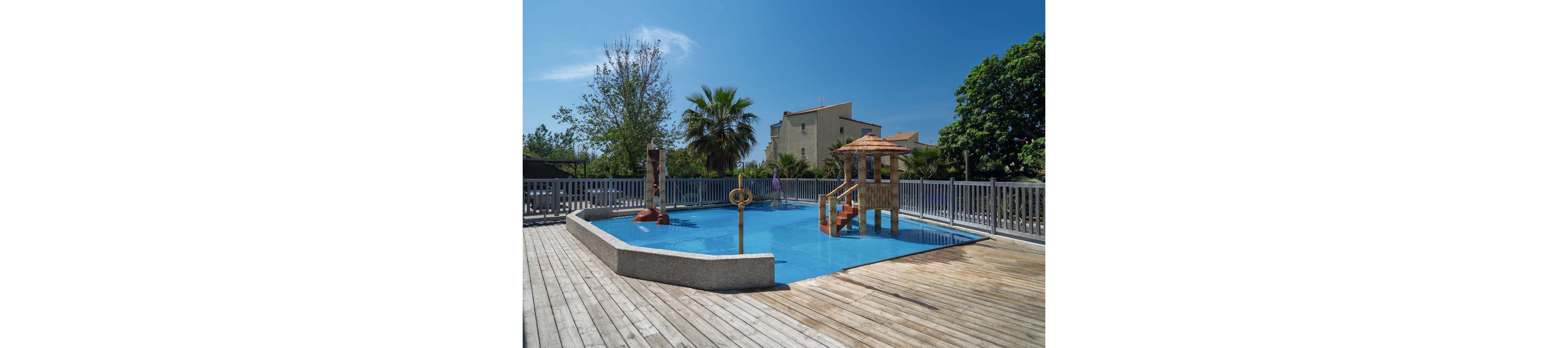 An outdoor shallow pool features a small wooden play structure with steps and a slide. The pool is surrounded by a wooden deck and a gray fence, creating a safe enclosed area. A person stands in the background near the fence, with residential buildings and lush green trees visible beyond. The bright blue sky and clear sunlight give the scene a warm, summery atmosphere.