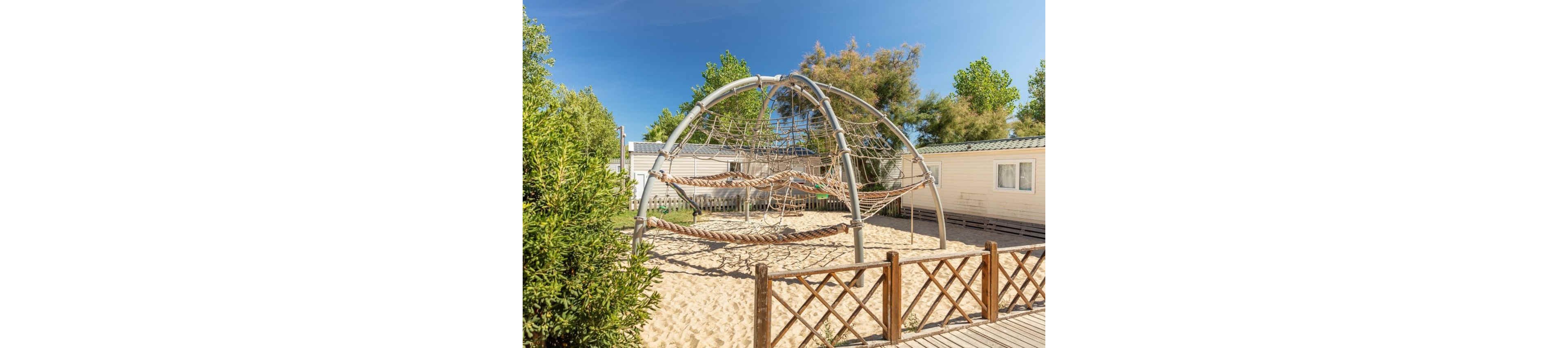An outdoor playground features a large metal climbing dome set in a sandy play area. The structure includes rope nets and suspended platforms for children to climb and balance on. Mobile home cabins and a wooden fence surround the playground, suggesting a holiday park or campground setting. Green trees and a clear blue sky create a bright, sunny atmosphere.