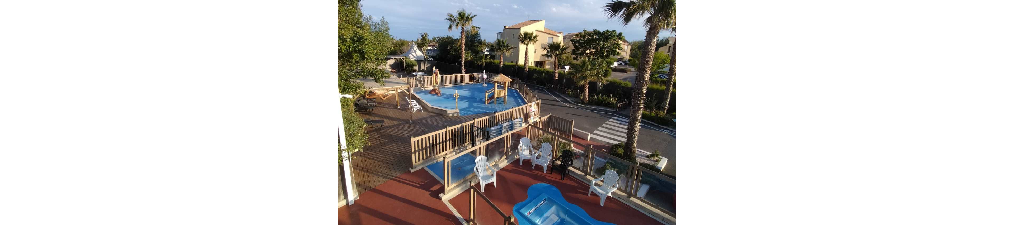 An elevated view shows an outdoor resort pool area surrounded by a wooden deck. Several white plastic lounge chairs are arranged around the pool, with a small blue wading pool in the foreground. Palm trees and landscaped greenery frame the scene, while residential-style buildings appear in the background. A quiet street with a crosswalk runs alongside the fenced pool complex, suggesting a relaxed vacation setting.
