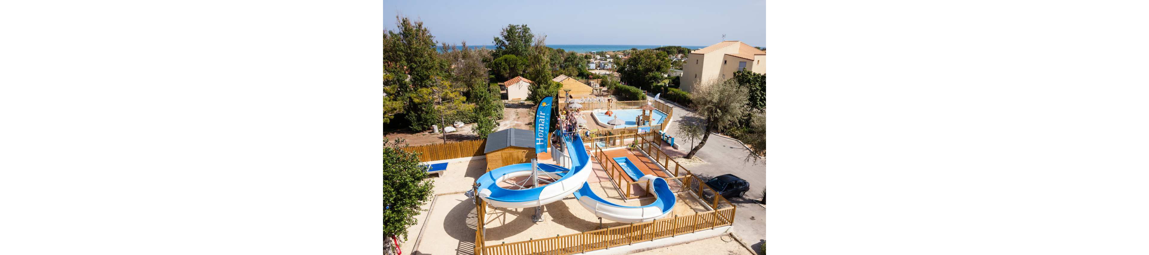 An aerial view shows a small outdoor water park with a blue and white spiral slide as the main attraction. The slide ends in a shallow pool, surrounded by wooden fencing and several smaller rectangular pools. A few people are visible enjoying the facilities under clear, sunny weather. Trees, buildings, and a distant view of the sea frame the recreational area, giving a relaxed holiday atmosphere. A tall blue vertical flag with the word "Homair" is prominently displayed near the slide.