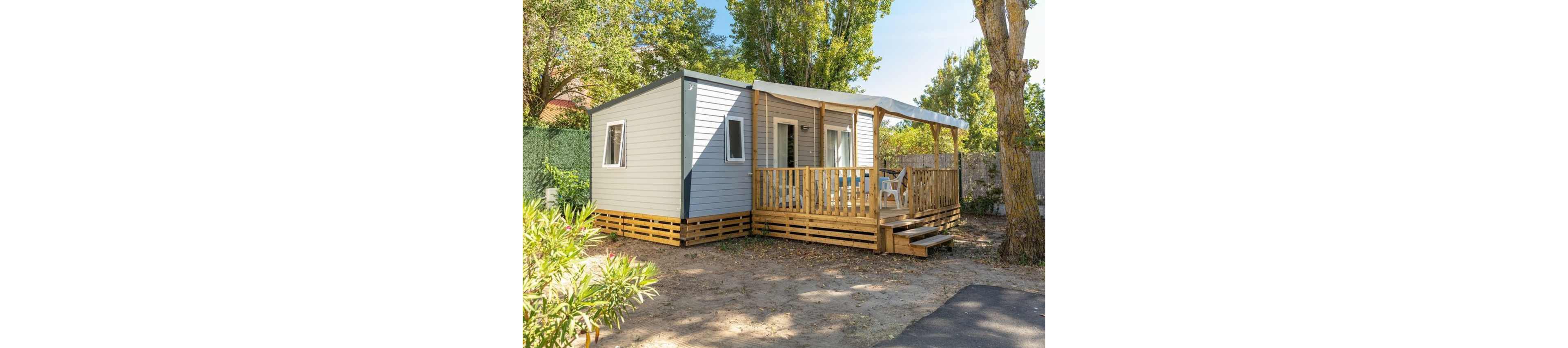 A compact mobile home with light gray siding and a wooden base stands in a sunny outdoor setting. The structure features a covered front terrace with a railing and a few plastic chairs, suggesting a relaxing seating area. Surrounding the home are trees, shrubs, and a mix of dirt and paved surfaces, indicating a campsite or holiday park environment. The overall scene feels calm and inviting, ideal for vacation or short-term stays.