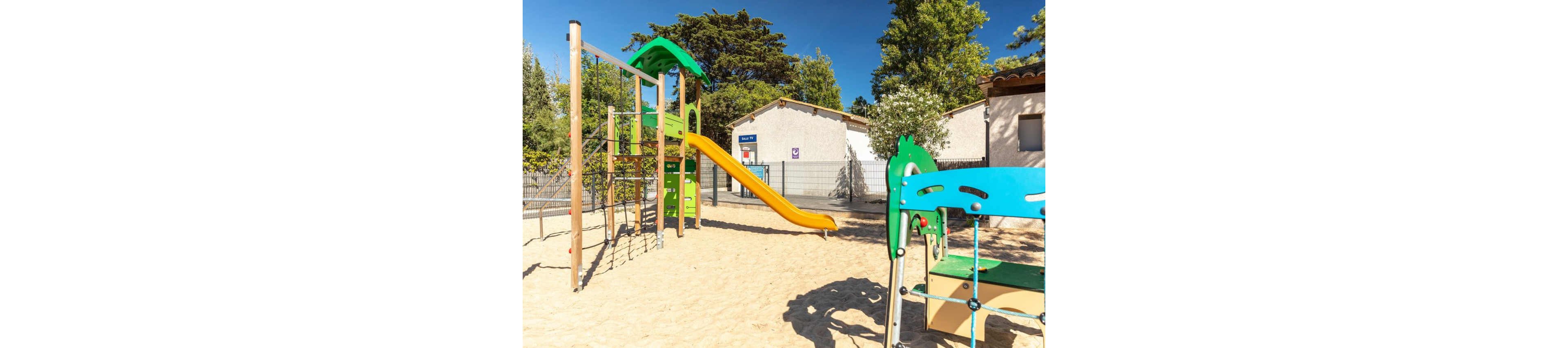 An empty outdoor playground features a tall climbing structure with a bright yellow slide set in a sandy play area. Colorful green and blue play equipment stands in the foreground, adding visual interest. The playground is surrounded by a metal fence and trees, with low white buildings in the background under a clear blue sky. Strong sunlight creates sharp shadows, giving the scene a warm and inviting atmosphere.