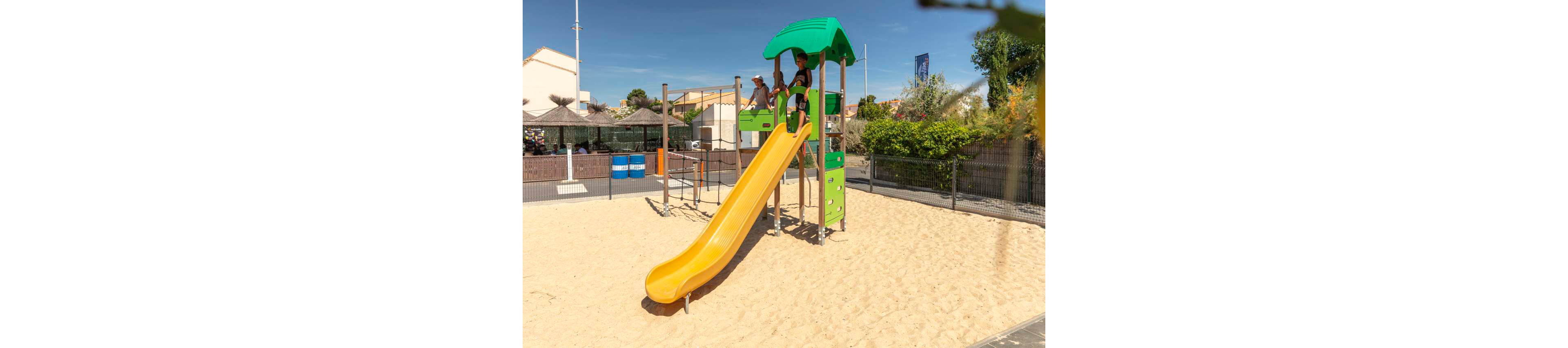 Two children play on a tall playground slide in a sunny outdoor park. The structure features a bright yellow slide with a green roof and panels, set over a large sand area. Fenced surroundings and nearby buildings suggest a secure recreational space in a residential or holiday setting. Clear blue skies and strong sunlight create a warm, cheerful atmosphere.