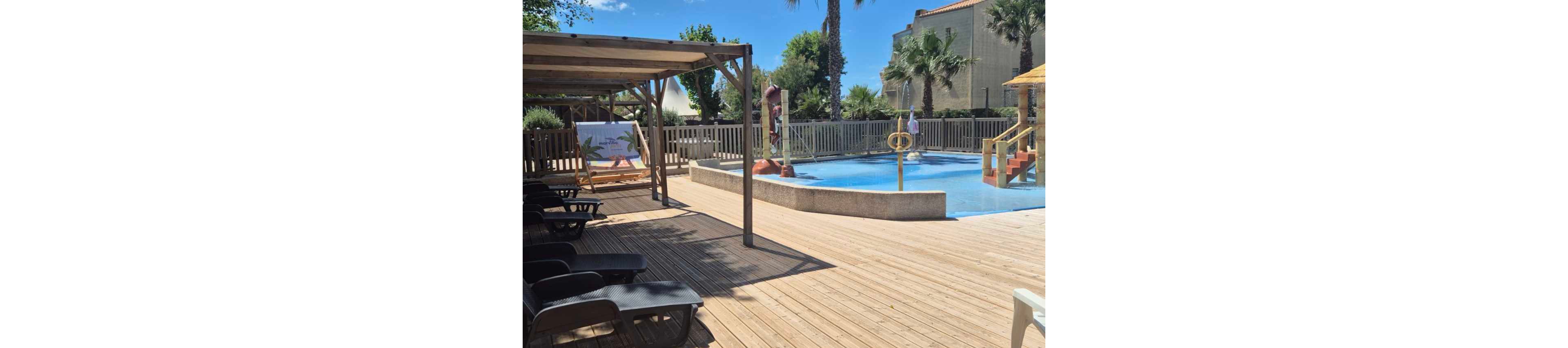 An outdoor swimming pool is surrounded by a wide wooden deck under a bright blue sky. Lounge chairs sit in the shade of a wooden pergola, offering a relaxing spot near the water. A small slide and playful water features are visible in the shallow pool area, enclosed by a fence and framed by palm trees. The scene suggests a quiet, family-friendly resort or campground pool on a sunny day.