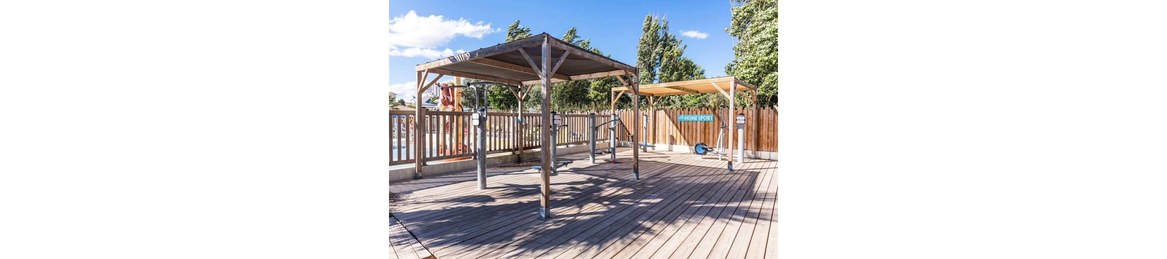 An outdoor fitness area features wooden pergola-style structures over exercise machines on a spacious deck. The equipment is arranged in rows, with metal posts and handles for various workouts. A wooden fence surrounds the space, and trees and a bright blue sky with scattered clouds are visible in the background. A sign on the fence reads "HOME SPORT," indicating the recreational nature of the area.