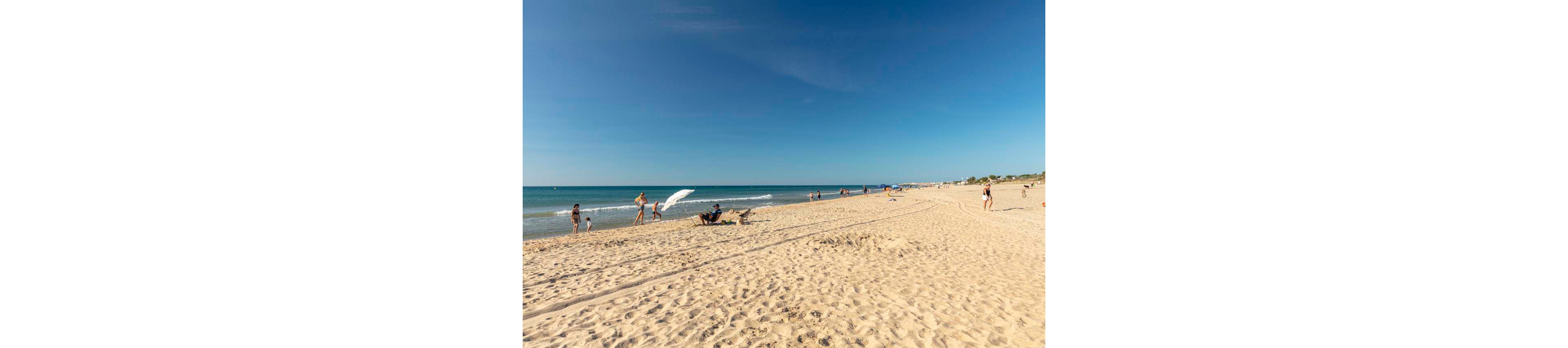 A wide sandy beach stretches along a calm blue sea under a clear sky. People are scattered along the shoreline, walking, sunbathing, and playing near the gentle waves. The bright sunlight casts soft shadows on the sand, emphasizing the warm, summery atmosphere. The horizon is low, giving a spacious, open feel to the coastal scene.