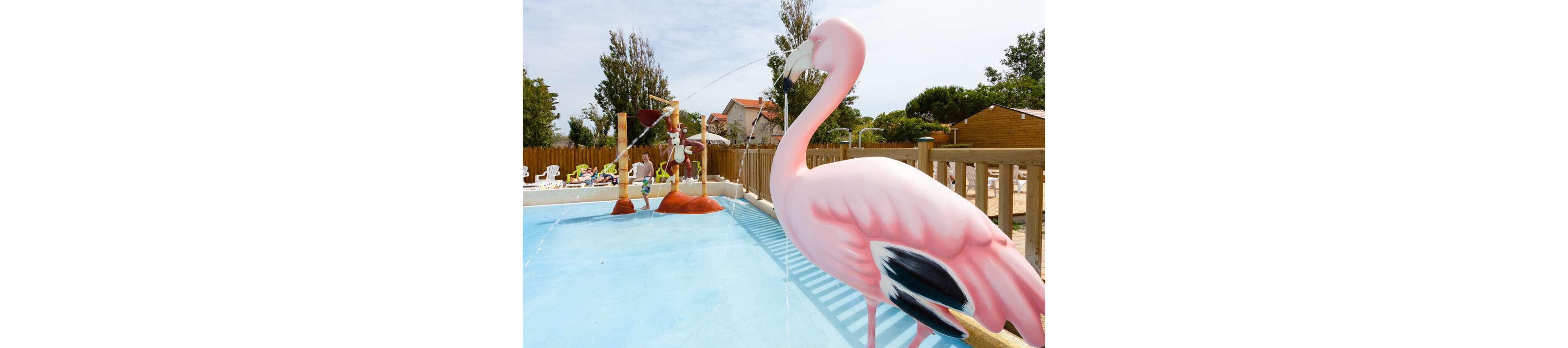 A large pink flamingo fountain stands in a shallow splash pool at an outdoor water play area. In the background, children play among additional bird-themed water features and small slides. The scene is set in a sunny recreational park with wooden fences, trees, and nearby buildings. The bright colors and playful sculptures create a fun, family-friendly atmosphere.