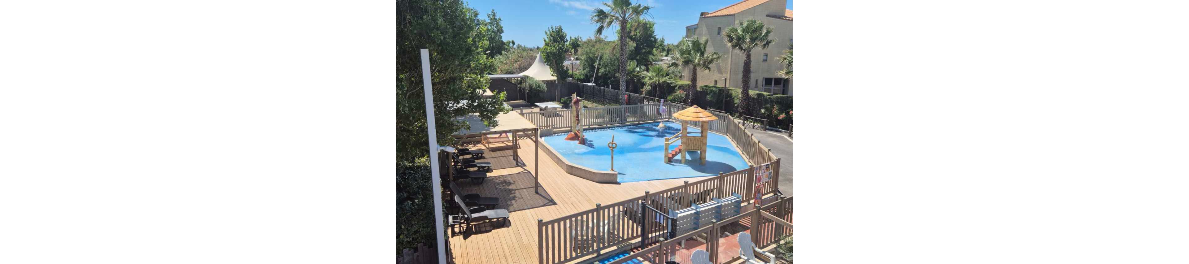 An elevated view shows a small splash pool in a resort setting surrounded by a wooden deck and safety fence. The shallow pool features a central water play structure with slides and spray elements designed for children. Sun loungers and shaded pergolas line the deck, offering relaxation areas for guests. Palm trees and nearby buildings frame the scene, creating a warm, vacation atmosphere under clear skies.