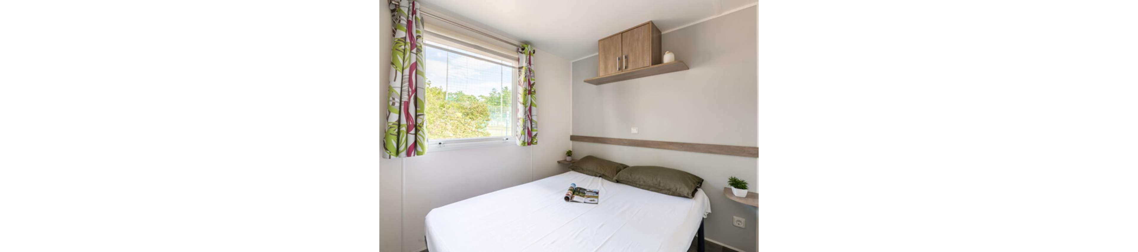 A small, minimalist bedroom features a neatly made double bed with white linens and two green pillows. Natural light streams in through a large window dressed with patterned curtains, revealing greenery outside. A wooden shelf with cabinets and small decorative plants adds warmth and storage above the bed. The overall space feels clean, bright, and modern, ideal for a compact apartment or vacation rental.